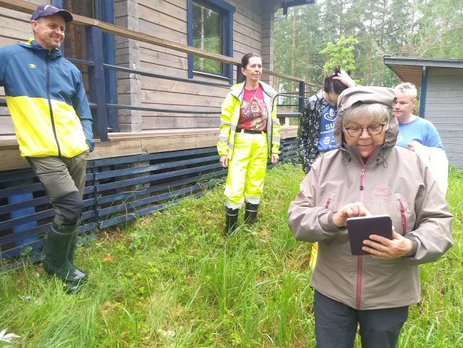 Anneli Helimo kaivoi tabletilta Matti Helimon videotervehdyksen, jossa Tampereen apulaispormestari esittelee kuivakäymälän sekä pesuvesien imeytyskentän toimintaperiaatetta. Taustalla Ari Toivari, Aicha Kivinummi, Katja Linnainmaa ja Marja-Liisa Suomalainen. huussikierros